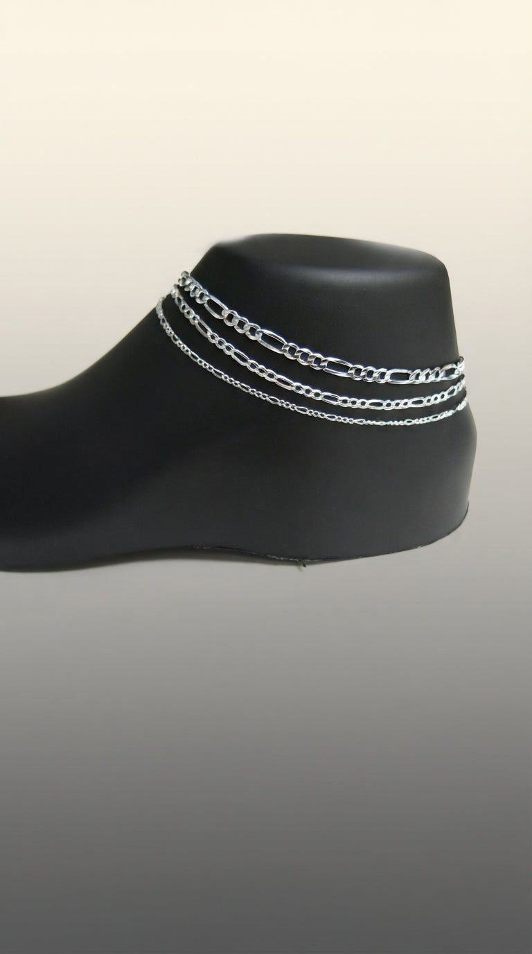 Close-up of a sterling silver Figaro link anklet worn on an ankle against a soft, neutral, out-of-focus background.